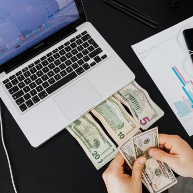 Person counting cash with laptop on desk, financial graphs visible.